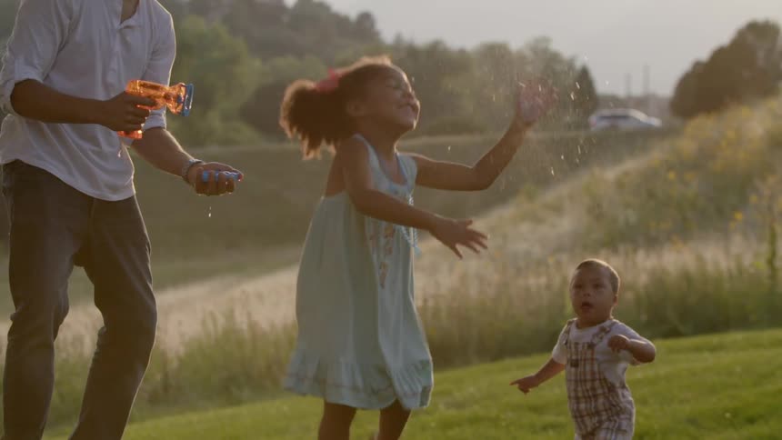 A father blows large bubbles for a young girl to pop in an open field