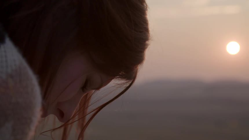 The wind blows a woman's hair as she looks down while the sun sets
