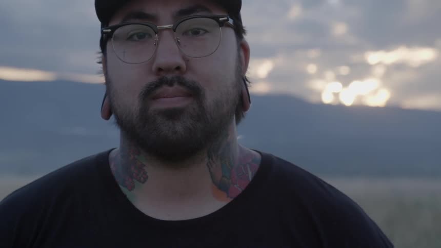 Bearded, spectacled man with a tattooed neck stationary with mountains and a cloudy sky in the background