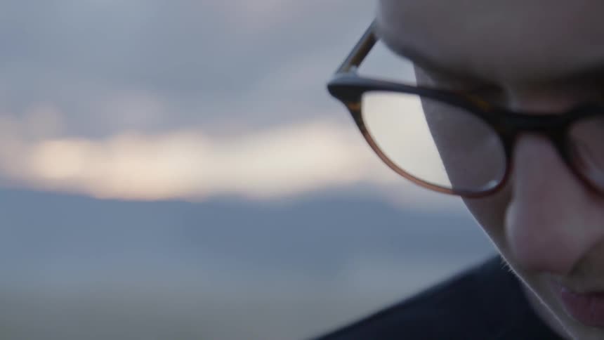 A man in glasses looks down with the sunset and mountains over his shoulder