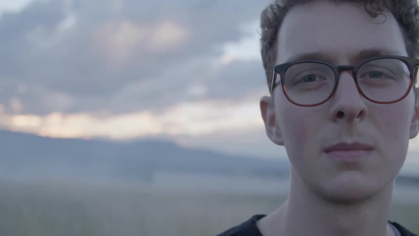 A young man wearing glasses standing under a cloudy sky with mountains in the background
