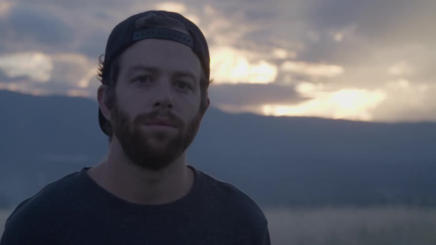 Bearded man wearing a baseball cap backwards standing in front of a cloudy sky