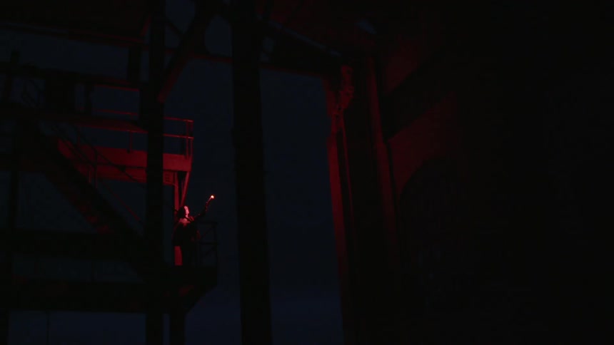 Someone stands in the darkness holding a red flair which illuminates buildings