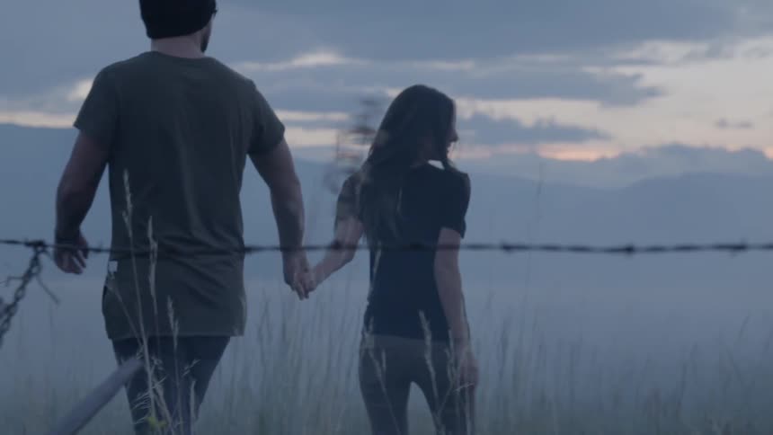 A man and woman hold hands and walk in a field at sunrise with mountains in the background