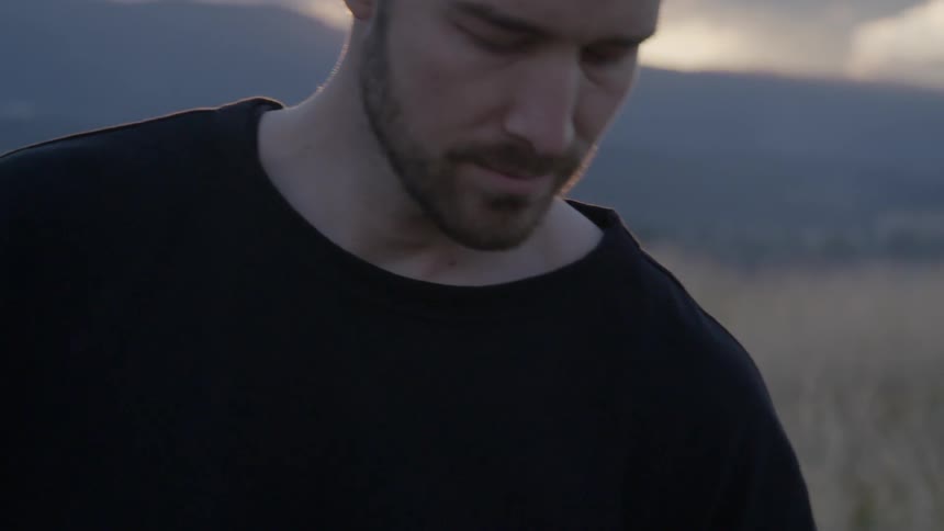 A sad looking man stands in natural setting as the sun rises behind him