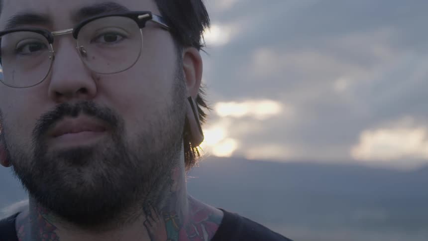 The face of a man with tattoos and earlobe plugs turns toward the camera