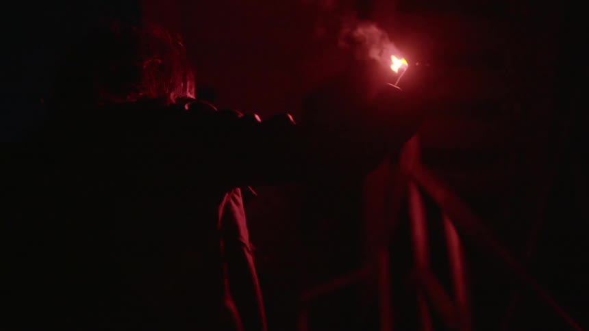 A man has a stick lit on fire and uses it like a torch to inspect a room with a staircase