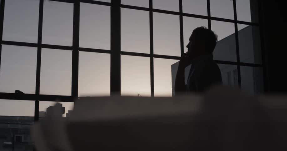 A man standing in an office looking out a window and talking on a phone