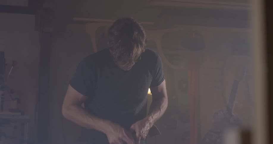 A man uses sandpaper on a piece of wood in a dark garage