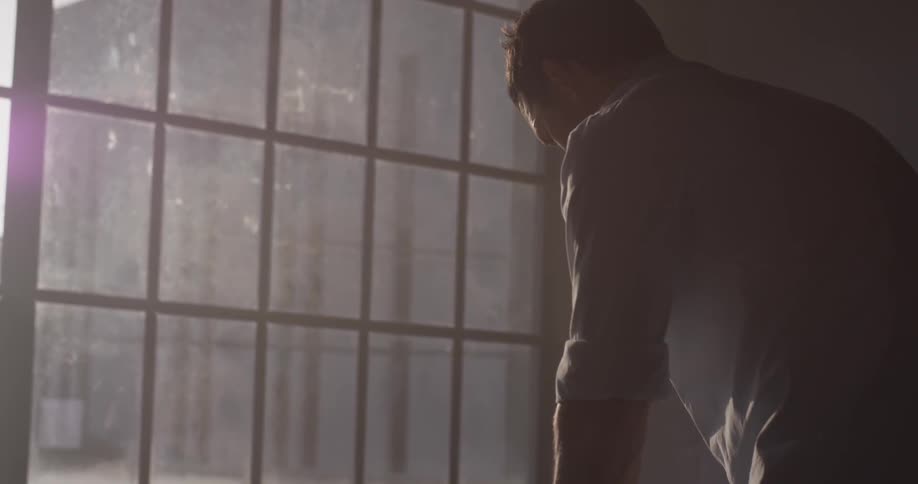 A man stands bent over in front of a window while he adjusts papers on a desk