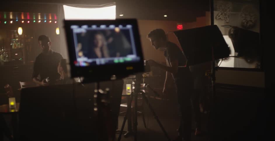 A monitor displays a scene as people make a video recording in the background