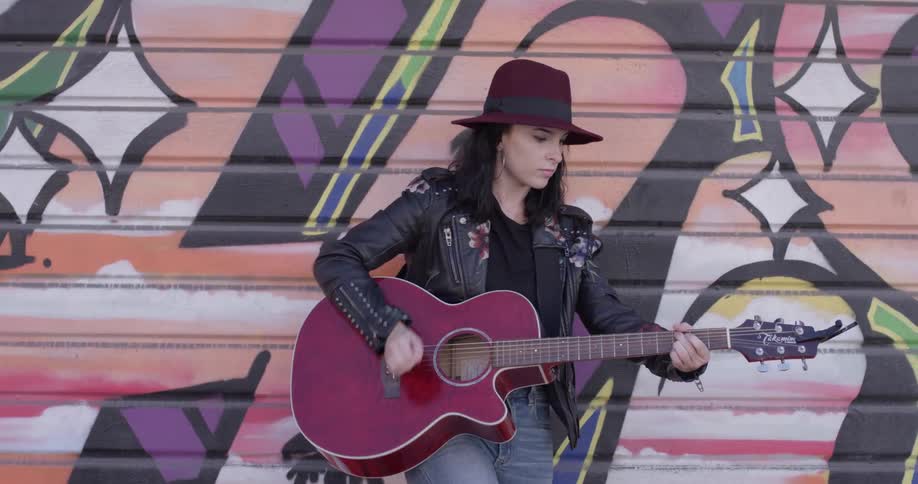 Woman in leather jacket and hat plays guitar in front of wall