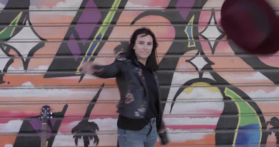 A woman poses then throws her hat in front of a graffitied wall