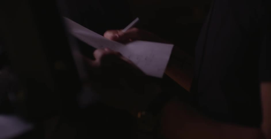 A man wearing headphones and looking at a computer screen, turns to accept some papers from a woman who walks up next to him
