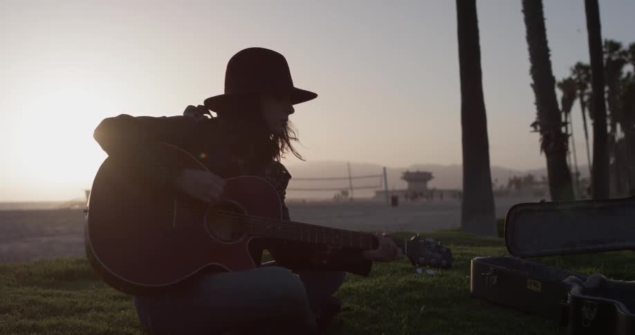 A woman plays guitar outside as the sun sets behind her