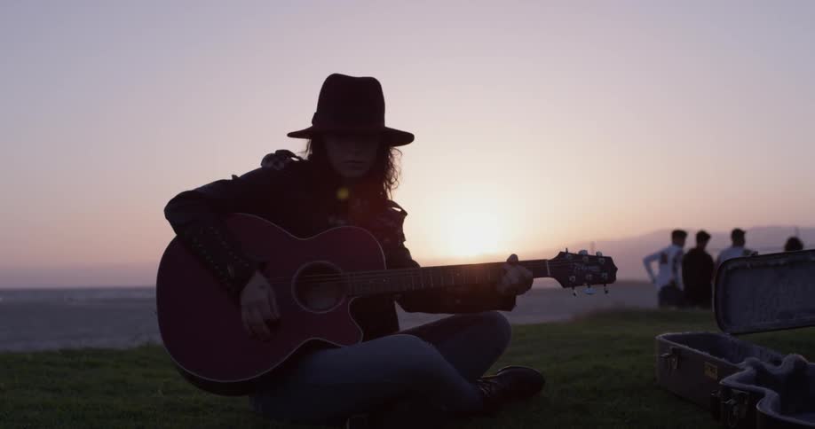 A person in a hat plays a guitar while sitting on grass