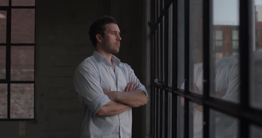 A man stands with his arms crossed while he looks out a window then walks away