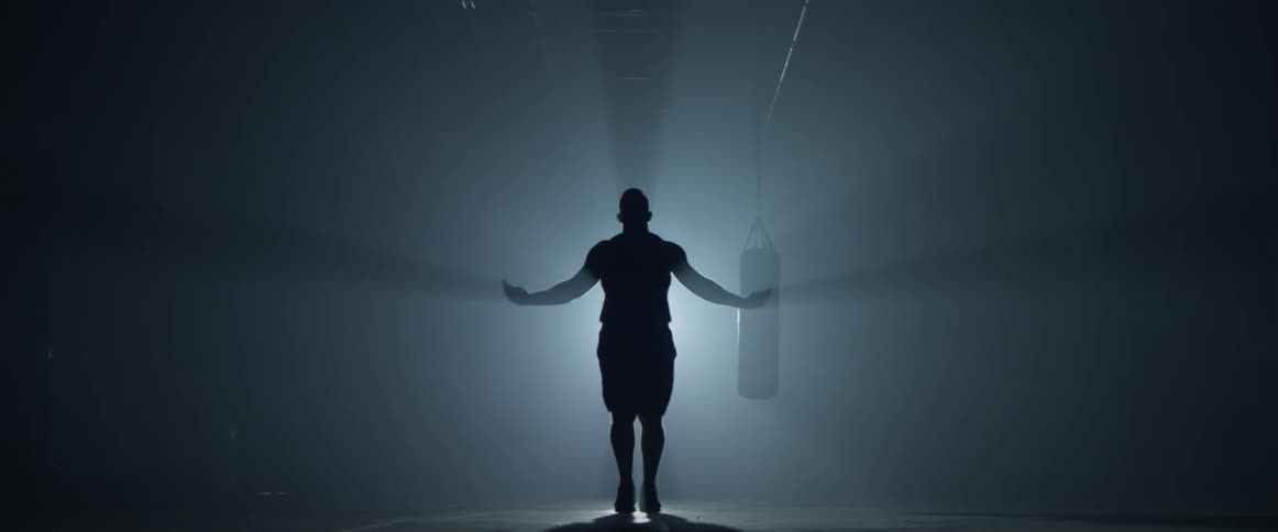 Man does jump rope in front of a spot light creating light beams from him and the punching bag behind him