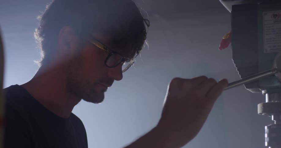 A man uses a drill press as light shines onto his back from a window