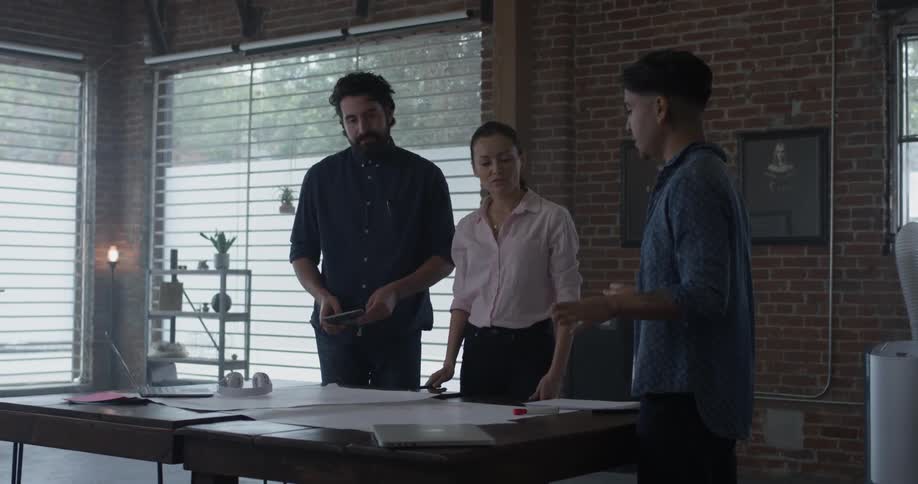 Three people hover over a desk and discuss things which are on paper and point to it