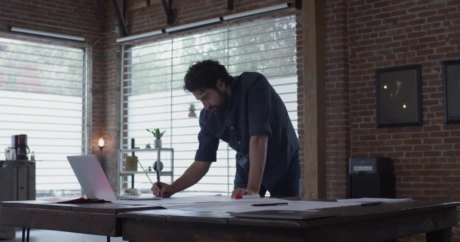 A man writes on some papers on a desk in front of a laptop