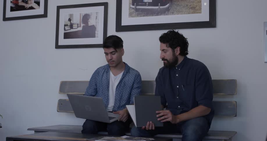 Two man sitting on a bench looking through a book and a laptop together