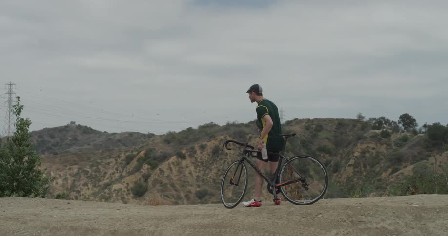 A man stands by his bike with his helmet in his arms while he takes a drink