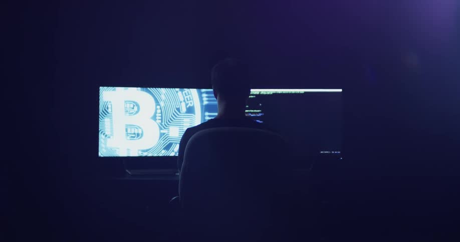 A man sits in front of two large illuminated monitors in the darkness