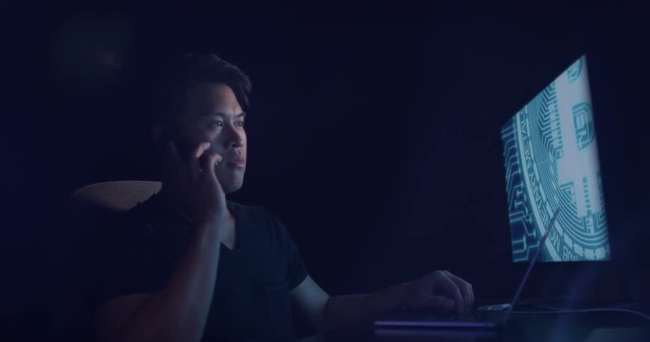 A man is on the phone in a dark room with a computer and laptop at a desk