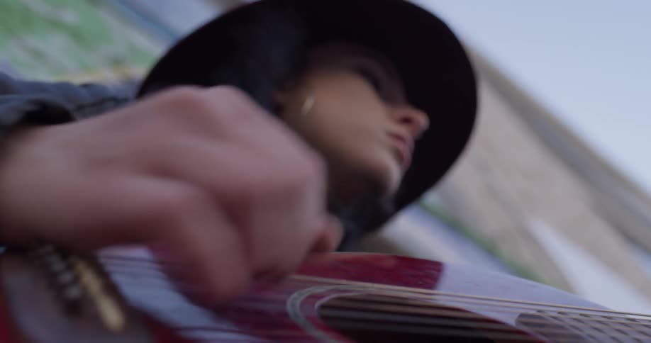 A woman strums and bangs on her guitar sitting up against a wall while wearing a jacket and hat in windy weather