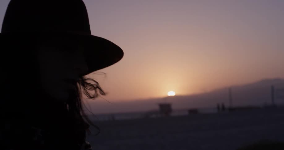 The silhouette of a person wearing a hat sways back and forth in front of a setting sun