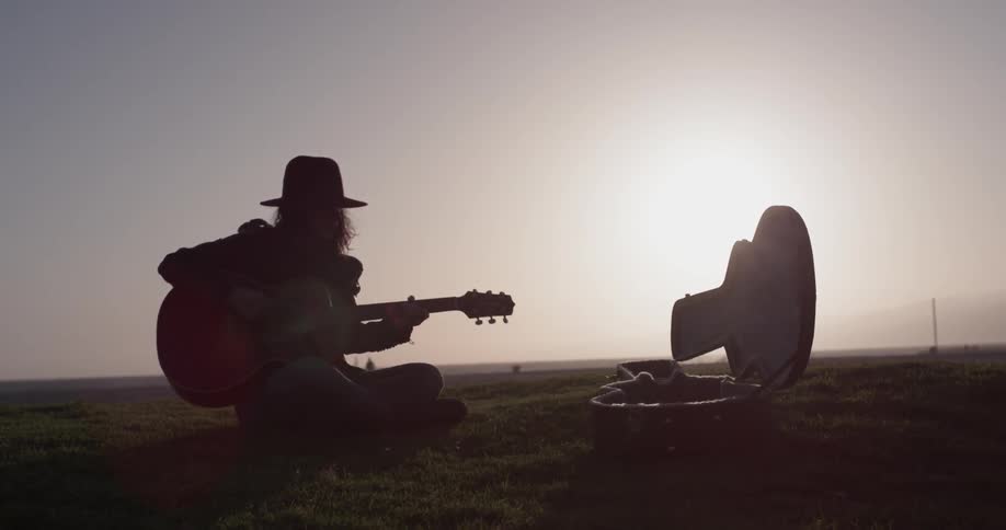 A woman sitting on a field and playing a guitar at dusk