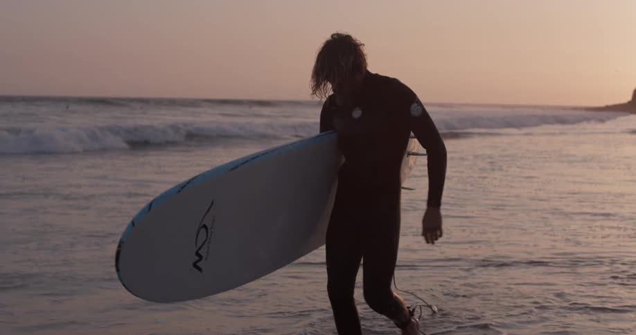 A surfer walks with his board under his arm by the ocean in the sunset