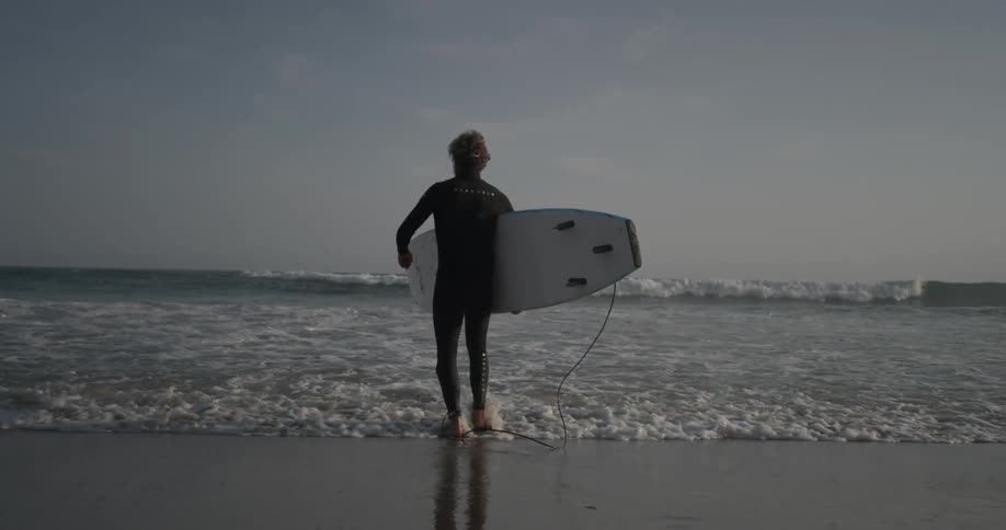 A man in a wet suit walks into the ocean carrying a surfboard