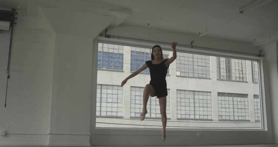 A woman in a black dress is leaping and doing the splits in the air at a dance studio in the city