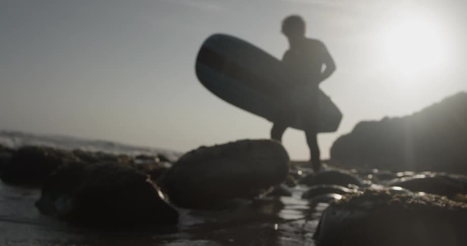 A man carrying a surfboard over some rocks near the beach