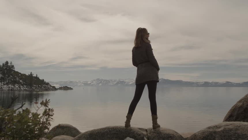 A woman stands on a rock and adjusts her sunglasses as she stares at a beautiful view of a lake