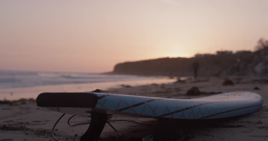 Surfboard lies on beach as man walks toward it at sunset