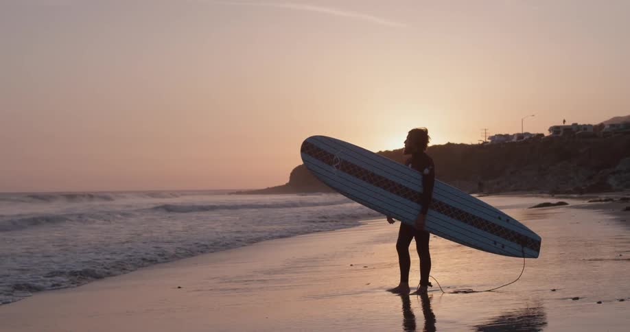 Surfer watches the ocean while holding his surfboard at sunset