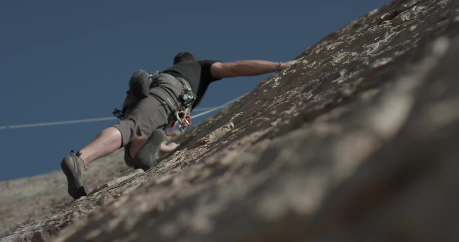 A man clings to the side of a cliff wearing a harness and moving sideways