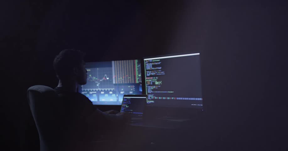 Man types on a laptop at a desk with two monitors in a dark room