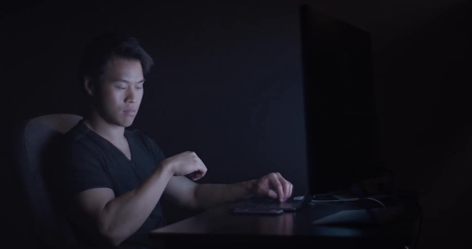 A man sits in the darkness on his phone lit only by his computer monitor