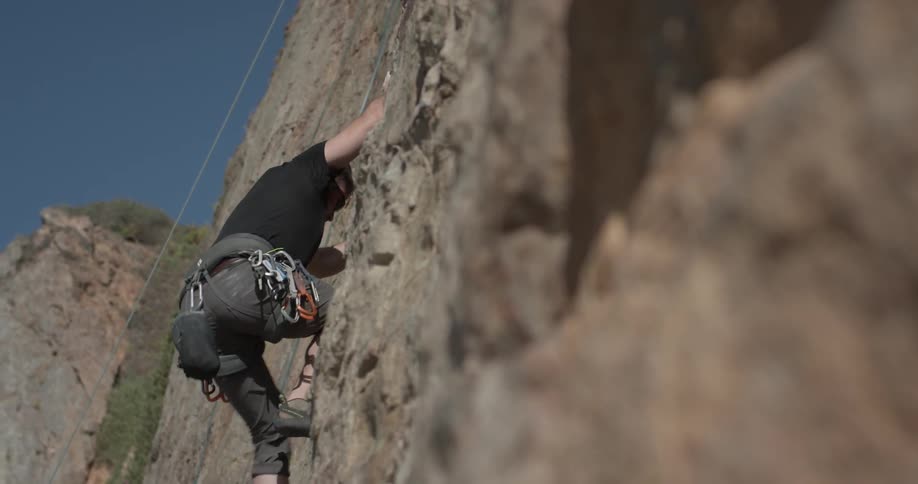 A rock climber scales a tall rock with blue skies above and climbing equipment