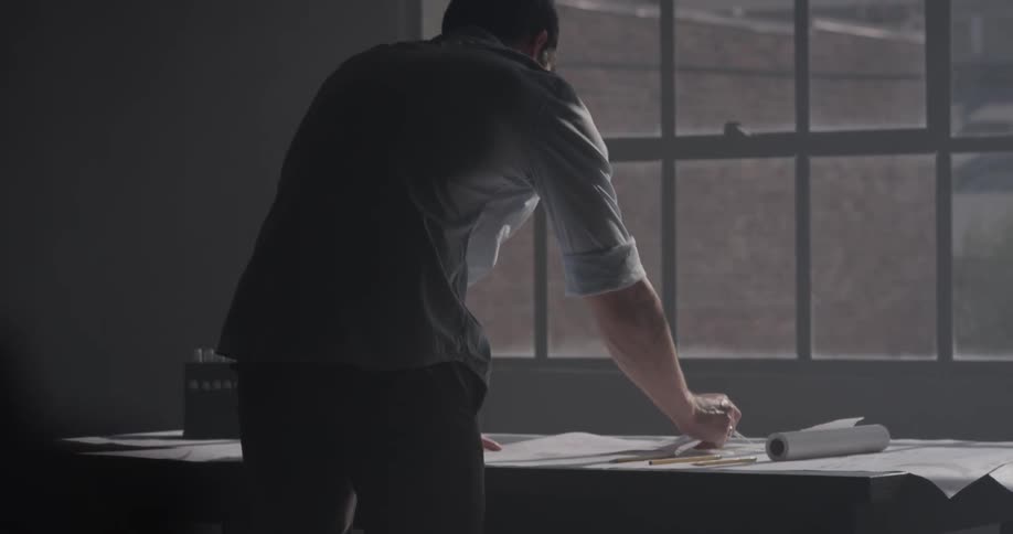 A man unrolls a paper on a desk in front of a window