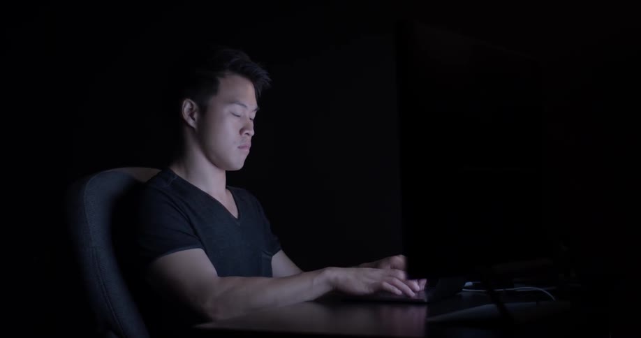 An asian man sits at a desk and presses keys on a keyboard