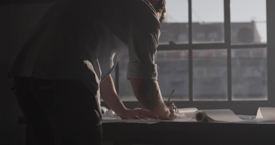 A man stands in front of a city window in the sunlight drawing on paper with a pencil