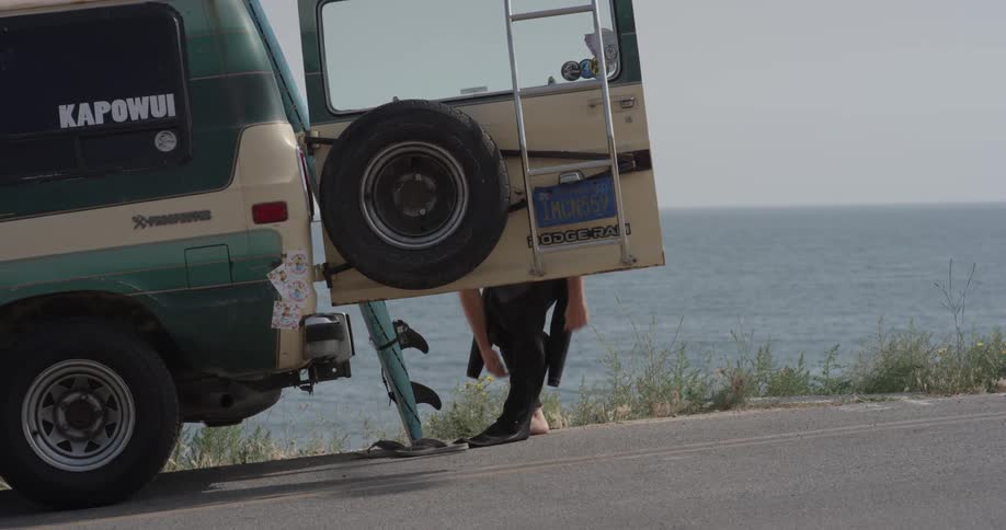 A person stepping into a wetsuit behind their open vehicle door