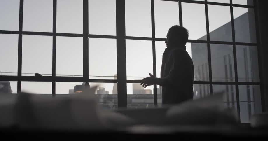 A man stands by a window and talks on a phone at dusk