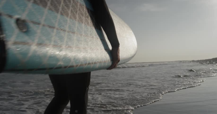 A man carries a surfboard with blue and black stripes wearing a wetsuit on a cloudy but bright sunny day at the oceanfront