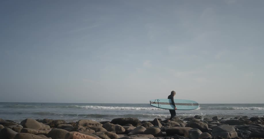 A man walking on some rocks along the coast carrying a surfboard